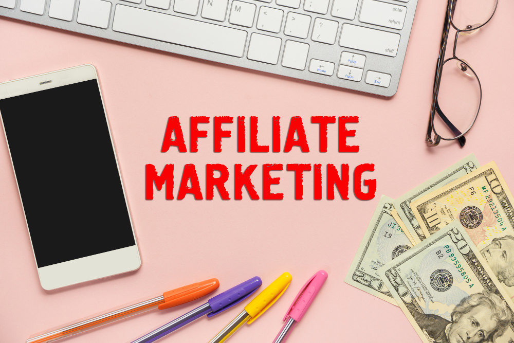 Keyboard, glasses, dollar bills, pens and a smartphone all surrounding Affiliate Marketing label in the center